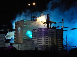 Cirque Bijou - a dancer stands on top of a set video mapped to look like a block of flats surrounded by atmospheric smoke