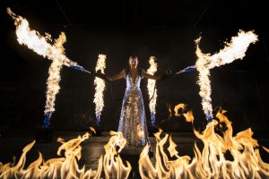 Cirque Bijou - A female artist wearing a glittery costume holds two massive flame throwers shooting fire, whilst fire explodes from the stage around her