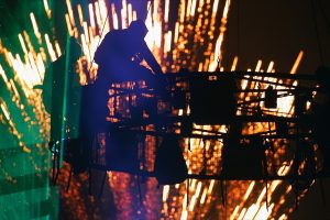 People sit on a circular steal structure in the air with fireworks in the background. 