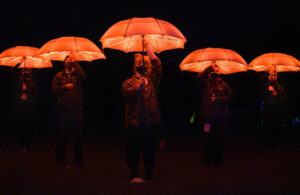 Community artists carry glowing red umbrellas to support WE MAKE EVENTS