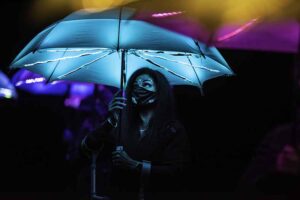 A community artist wearing a facemask holds aloft a glowing blue umbrella