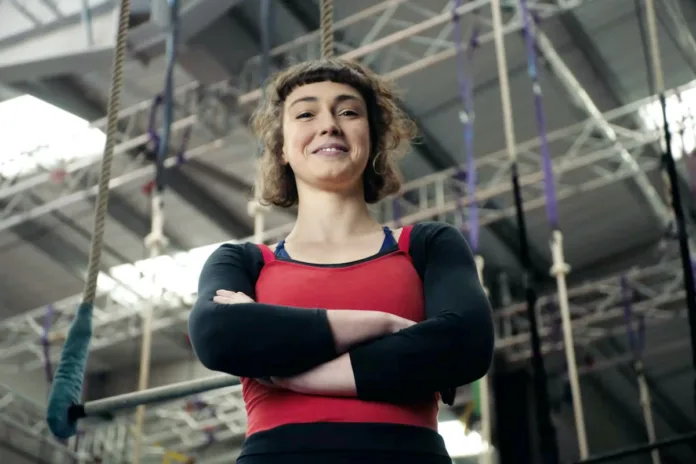 Tilly Lee Kronick stands with her arms crossed smiling down at the camera, with truss and rigging in the background