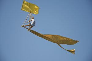 A woman poses in the air against a beautiful blue sky, with yellow silks flying around her