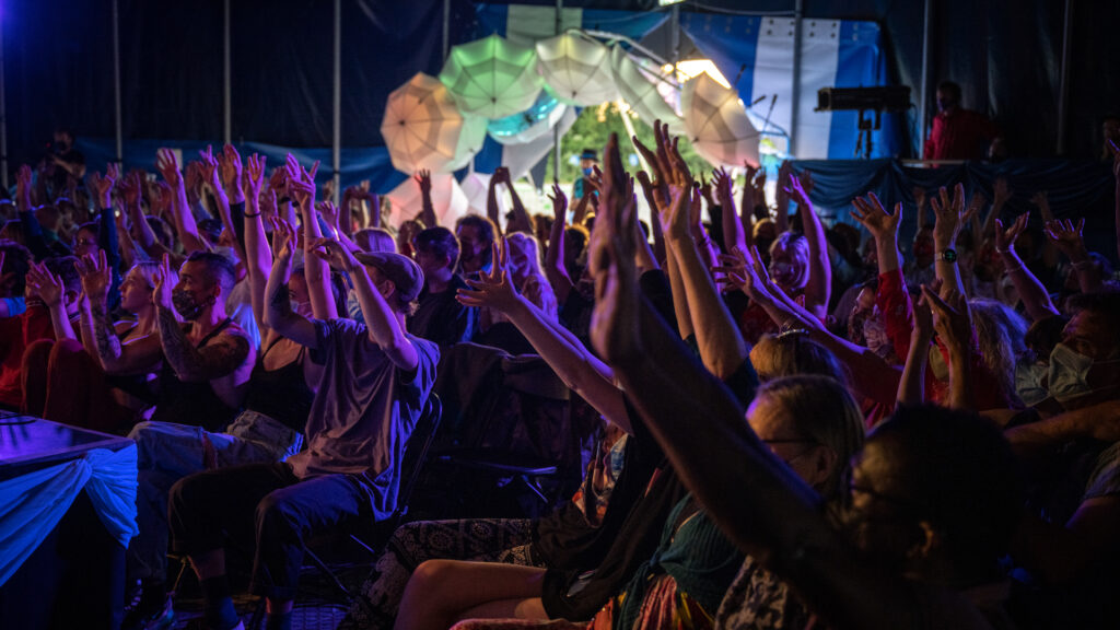 An audience wearing masks have their hands up in their air, facing towards a stage off screen.  They are in a Big Top tent with an entrance way  with an arch of colourfully lit umbrellas