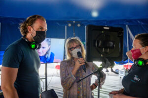 Three people in a rehearsal room wearing masks gather around a screen, one with a microphone