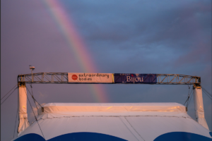 A rainbow against an atmospheric sky falls down on to the top of a Big Top, with banners on the rigging reading 'Cirque Bijou' and 'Extraordinary Bodies'