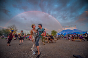 A male and female partygoer pose in glittery clothing, big top tent behind them and rainbow overhead against a dramatic blue and cloudy sky. They are distanced from a scattering of audience members around them.