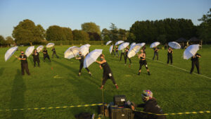 A field of distanced dancers hold white umbrellas up in a synchronised movement, green grass and blue sky