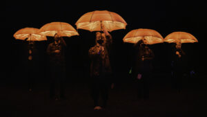 A phalanx of people advance holding glowing red umbrellas above their heads against a dark background. Their mouths and noses are hidden behind masks