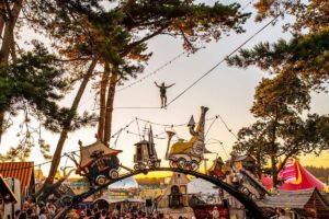 A man balances on a high wire, amongst the trees and above some colourful, ramshackle sets.