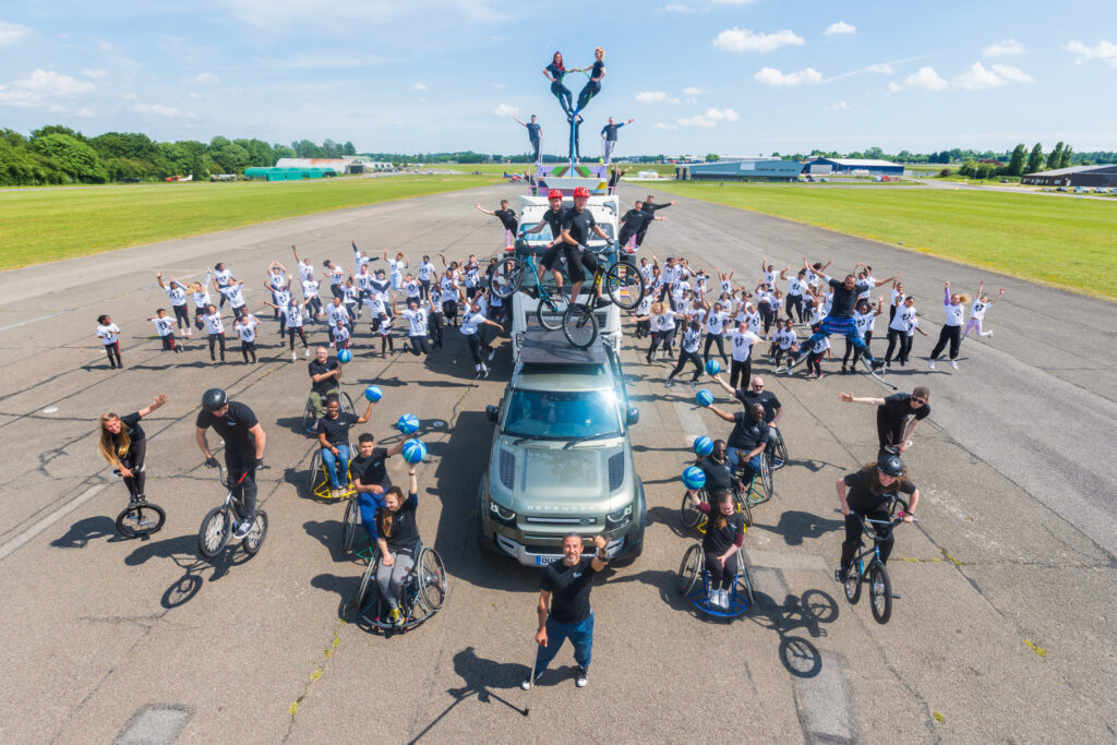 A group shot on an airport runway showing vehicles, dancers, wheelchair users, bicycles, unicycles, aerialists on top of the truck cab - around 100 people in all, everybody waving 