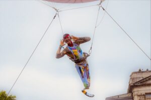 Close-up on aerial artist. An amputee from below the left knee, he is wearing a blade. He wears a colourful costume and flamboyant makeup. He holds his hands to his ears as though listening (for applause)