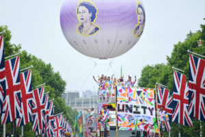 Union jacks and leafy trees line the Mall. A giant spherical helium balloon, printed with an image of the young Queen Elizabeth II floats above a truck covered in people waving, wearing brightly coloured outfits. In the foreground a colourful banner reads the word UNITY