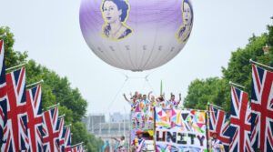 The Mall is lined with Union Jack Flags and leafy green trees. Above, a giant spherical helium balloon floats, printed with an image of a young Queen Elizabeth. An aerialist is suspended below the balloon. Ahead of this a colourful truck can be seen, with a number of people waving on top, dressed in brightly coloured clothes. To the front, a large colourful banner reads UNITY.