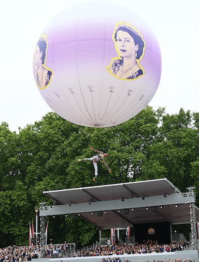 A huge spherical helium balloon, printed with an image of a young Queen Elizabeth II, flies above a crowded grandstand, an aerialist suspended underneath.