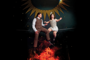 A man and woman sit together on a tightwire. The man looks longinngly at the woman, grapsing her hand, whilst the woman looks off in the distance with a smile on her face. Embers of a fire reach up from the bottom of the image.