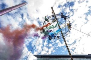 Colourful smoke effect against a blue sky with mackerel clouds, with a ring-shaped structure suspended from a crane, on which artists perch in colourful costumes