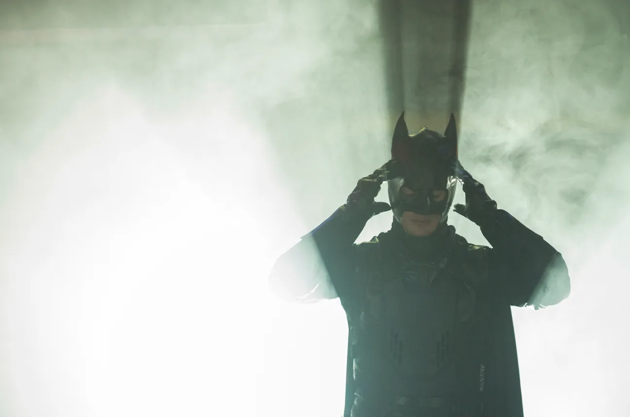 An actor dressed as batman adjusts his cap in front of green smoke