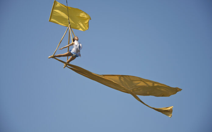 An aerialist poses dramatically against a clear blue sky - yellow flags fly around her