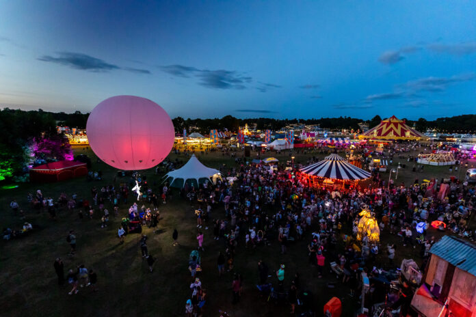 Camp Bestival Birdseye view of an aerial performer attached to a large pink ballon. There are big tops in the background and large crowds gathered around the big tops.