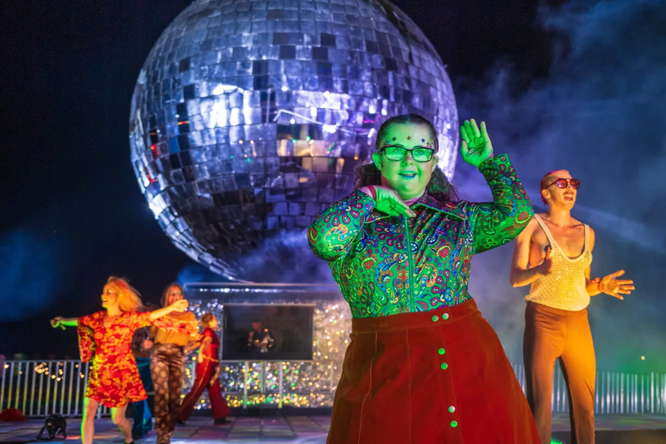 Performers in bright disco clothing stand in front of the giant silver disco ball at Camp bestival.
