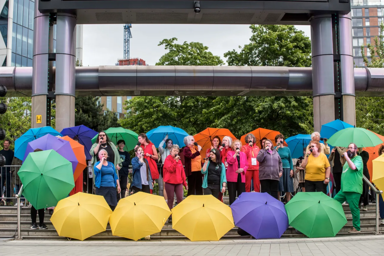 A choir dressed colourfully sing with mics and colourful umbrellas surround them.