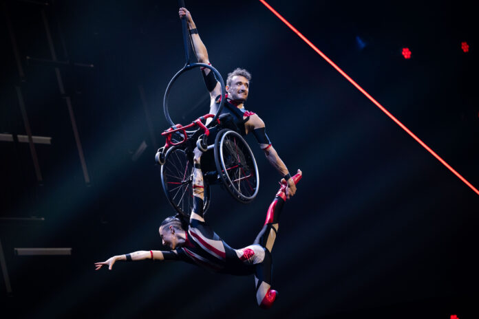 Two aerialists in red and black costumes are suspended in the air against a dark background. Jonny is sitting in a wheelchair and hanging by one hand from a strop. Charlotte, below, holds the bottom of Jonny's wheelchair and strikes a graceful pose supported by Jonny.
