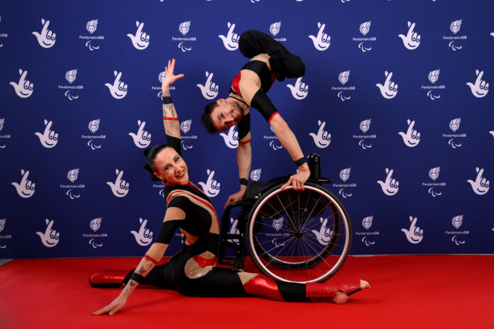 Jonny and Charlotte pose and smile for the camera on the red carpet, in front of a National Lottery and ParalympicsGB branded backdrop. In black and red costumes, Charlotte is doing the splits and Jonny is standing on his hands atop his wheelchair.