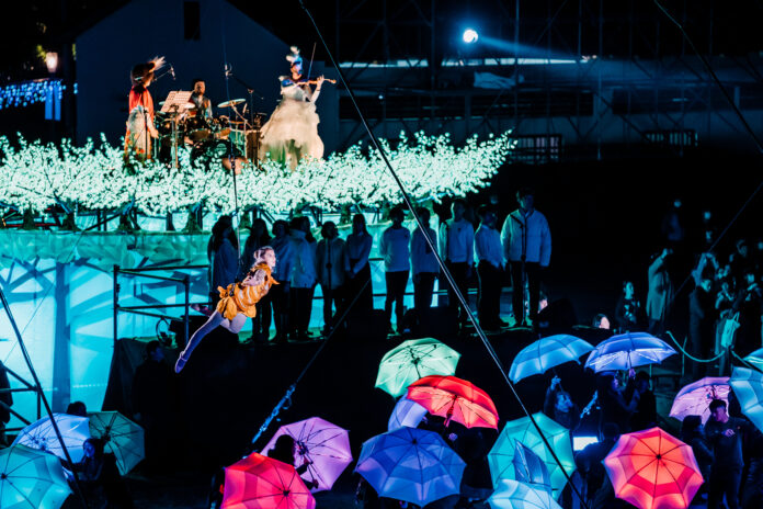 Aerialist in light up dress flies over performers holding light up umbrellas at night