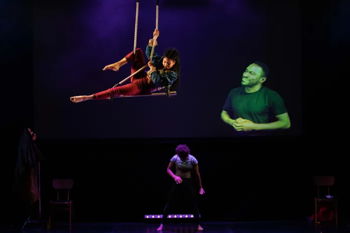 An aerialist performs on a trapeze whilst a BSL interpreter and performer below are also in the image