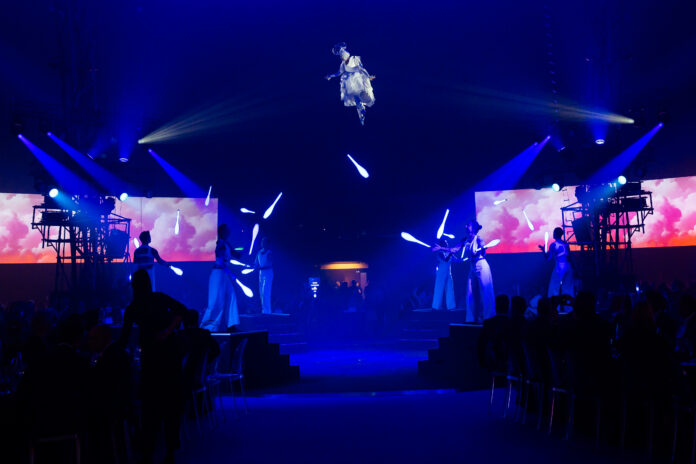 An aerialist hangs suspended from the ceiling with spotlights all around her, and jugglers performing beneath her
