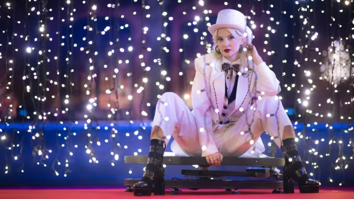 An artist sits in a white suit and top hat, with against a backdrop of fairylights