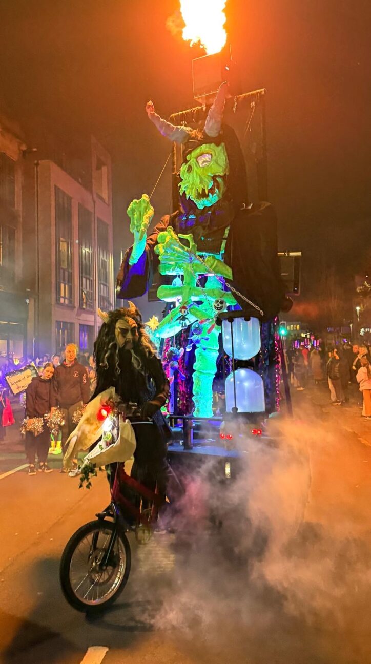 Demon on a bike with large skeleton puppet on the back