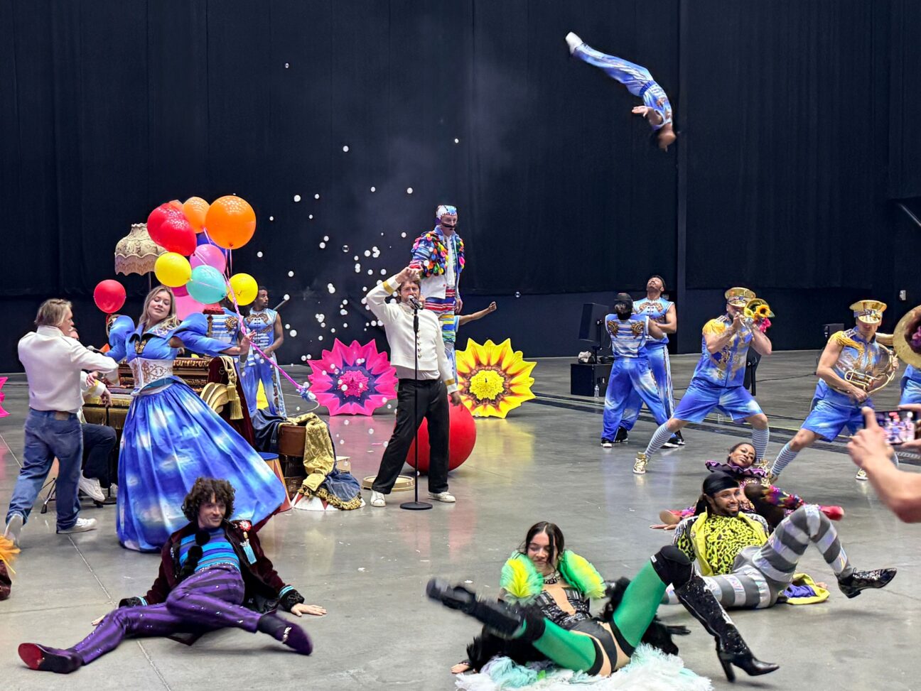 Photo of rehearsal with Take That band surrounded by circus performers all dancing with one performer doing a backflip high in the air