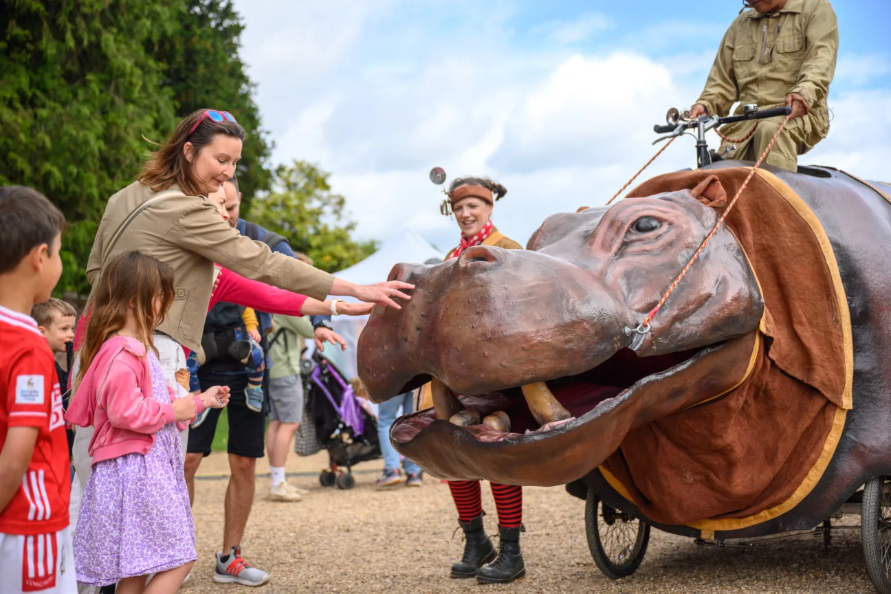 Some children interacting with a large mechanical hippo with a safari performer riding on it.